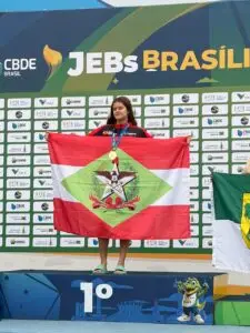 Estudante-atleta catarinense com medalha e bandeira de Santa Catarina no pódio dos JEBs Brasil